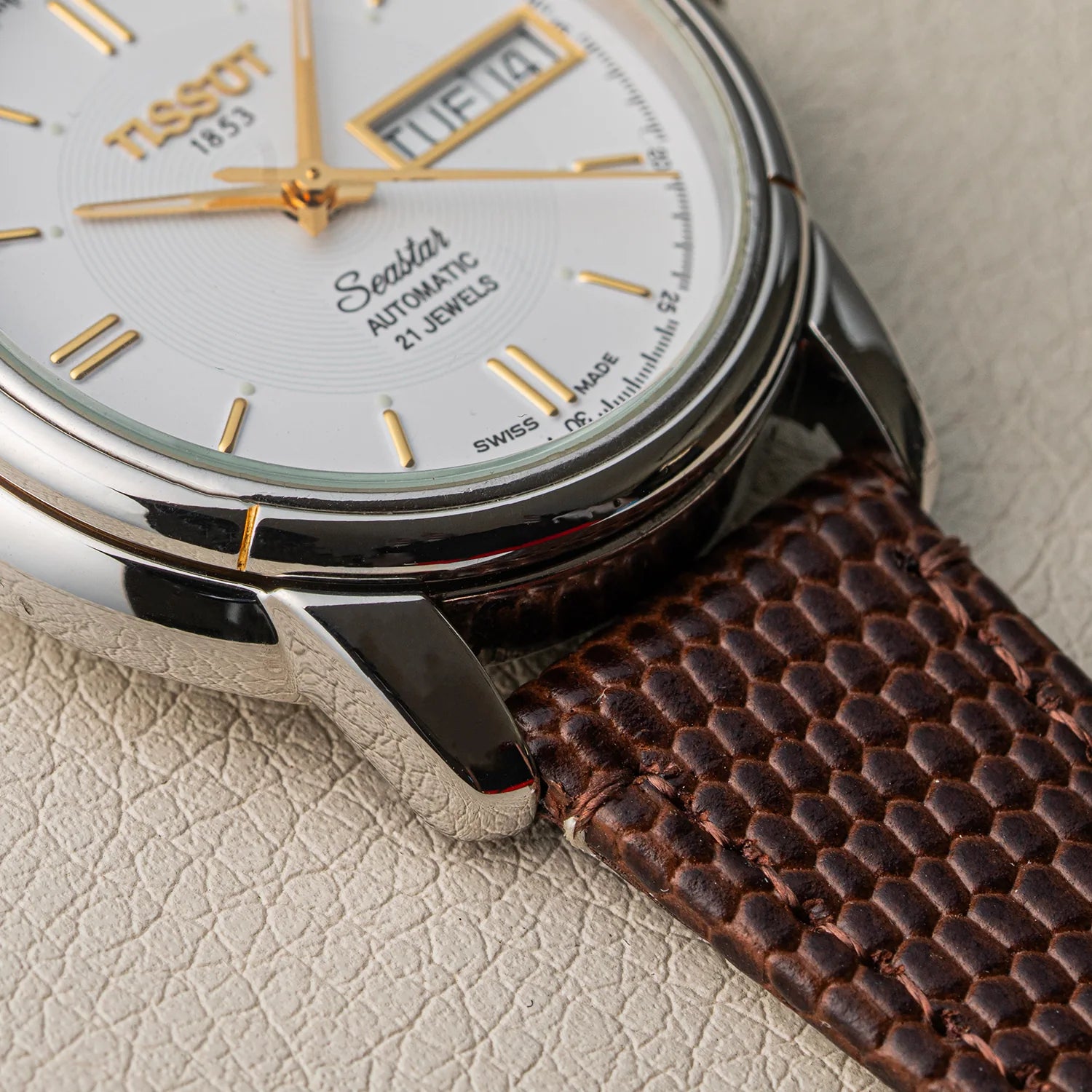 Close-up of a Tissot Seastar ETA 2846  automatic watch with a silver-toned case, white dial, gold hands and markers, and a brown textured leather strap on a light beige textured surface.