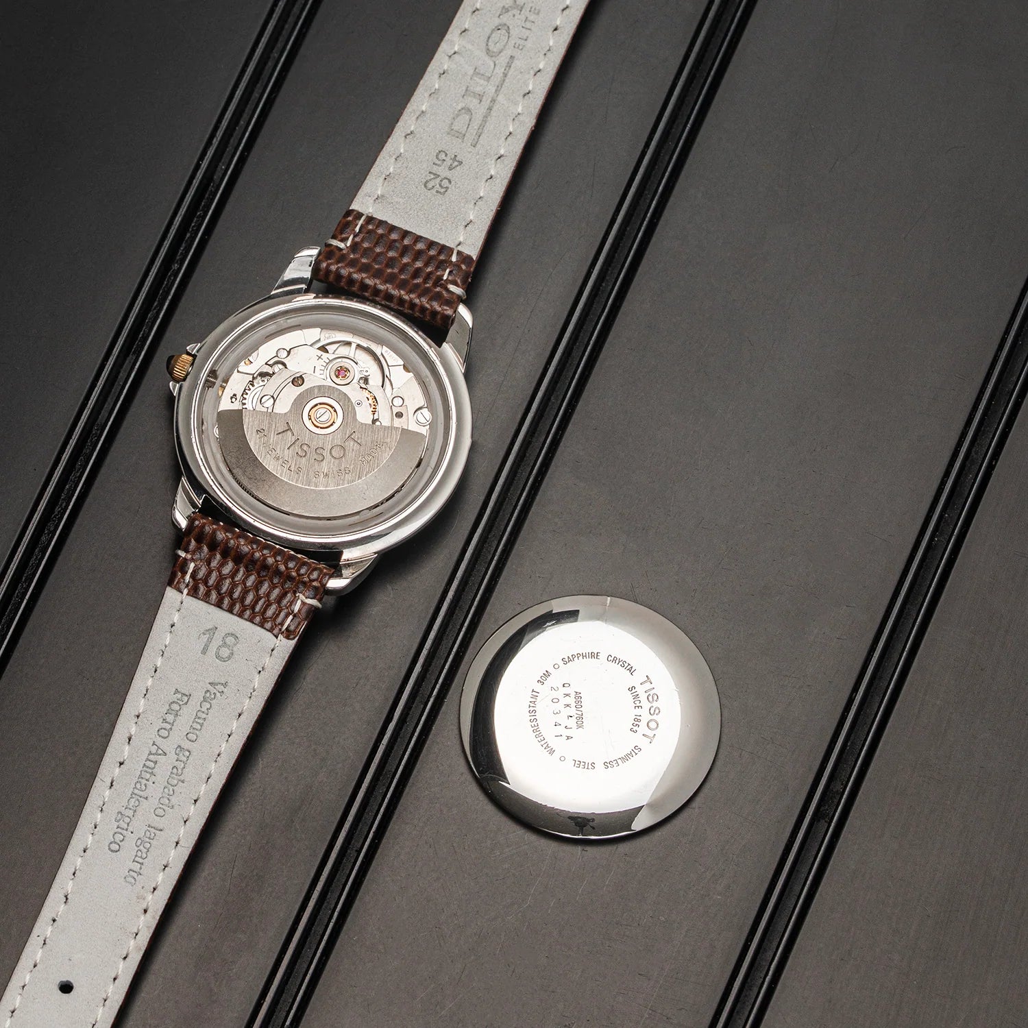 A close-up of a wristwatch Tissot Seastar ETA 2846  shown from the back, with the case removed and placed beside it on a dark textured surface. The watch has a silver-toned case, a brown woven strap, and a white dial with printed text.