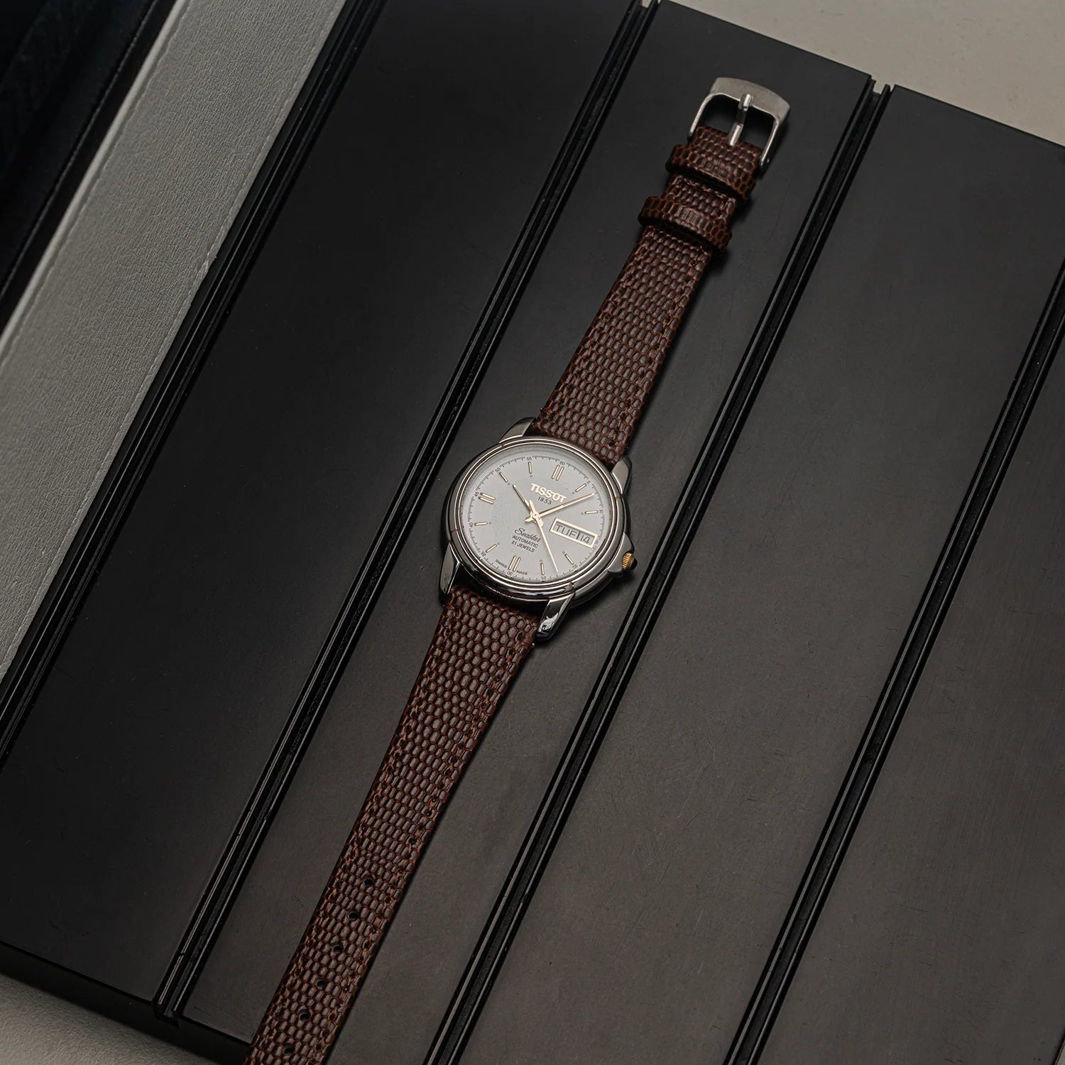 A wristwatch Tissot Seastar ETA 2846  with a brown woven fabric strap and a silver-toned dial, laid diagonally on a dark textured surface.