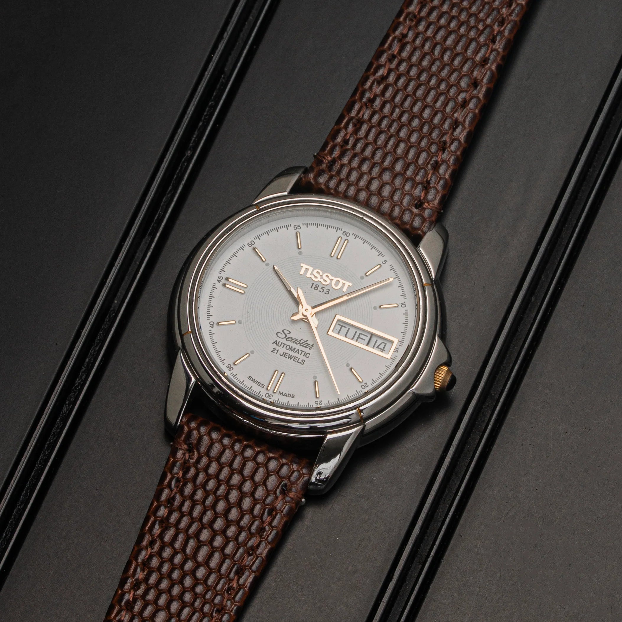 A Tissot Seastar ETA 2846  watch with a silver-toned case, light gray dial, and textured brown leather strap, displayed diagonally on a dark textured background.