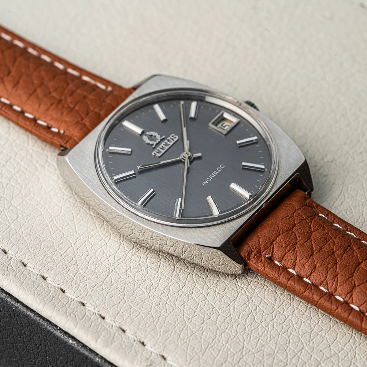 A close-up of a vintage-style Titus Geneve, Ref. 9379, Cal. FHF 96-4, Men’s Manual Wind, 1972 wristwatch with a square silver-tone case, dark gray dial, and brown leather strap with white stitching, resting on a textured off-white surface.