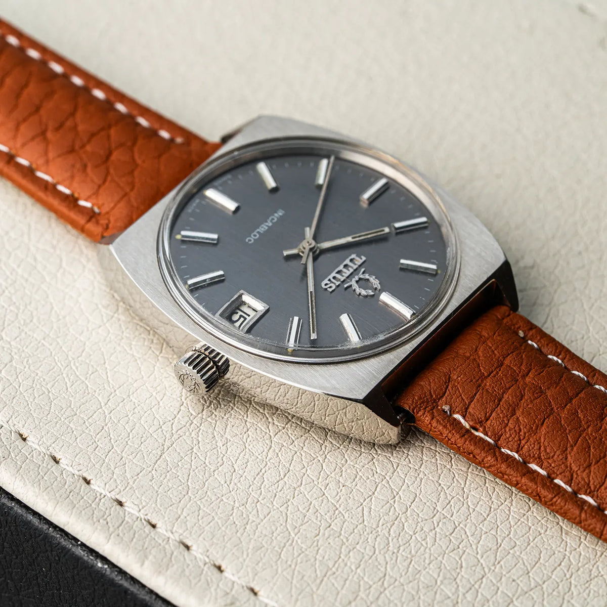 A vintage-style wristwatch Titus Geneve, Ref. 9379, Cal. FHF 96-4, Men’s Manual Wind, 1972 with a silver-toned case, dark gray dial, and brown textured leather strap, photographed on a light cream textured surface.