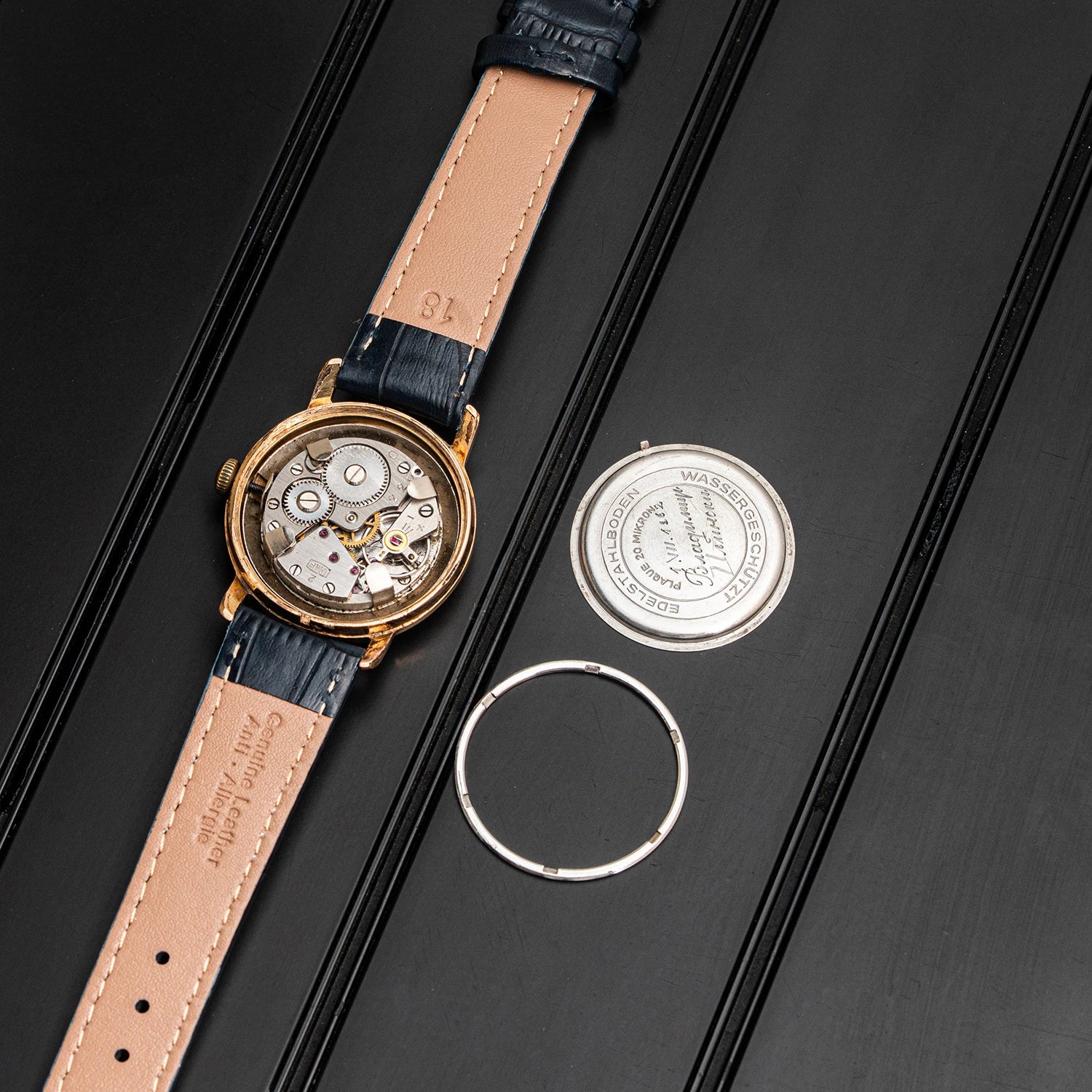 A luxury watch with a gold-toned case, a tan leather strap, and a dark blue alligator leather loop, displayed alongside its silver metal case back and ring on a black textured background.