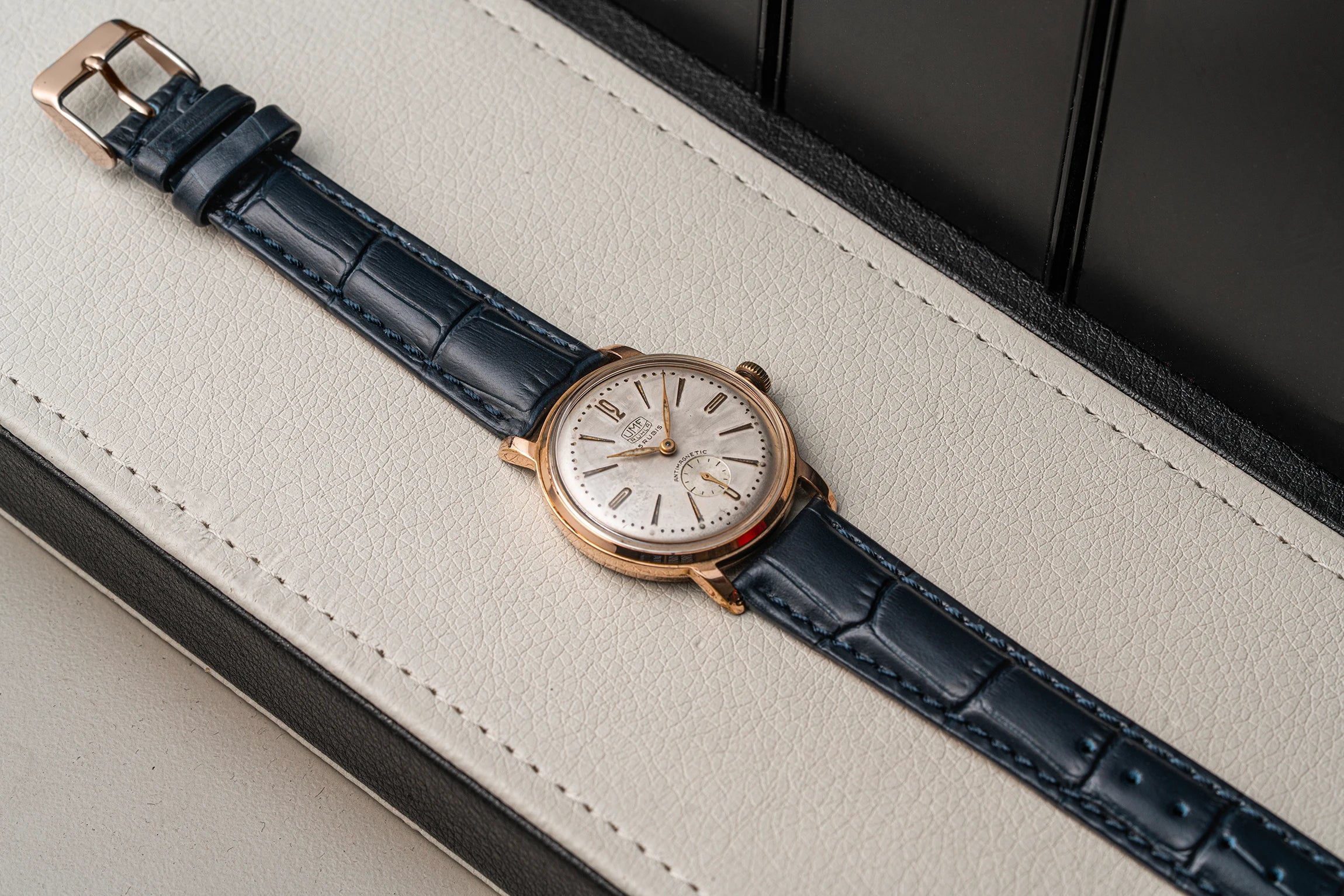 A vintage-style UMF Ruhla, Cal. UMF 2 wristwatch with a rose gold case and a dark blue alligator leather strap, resting diagonally on a cream-colored leather surface.