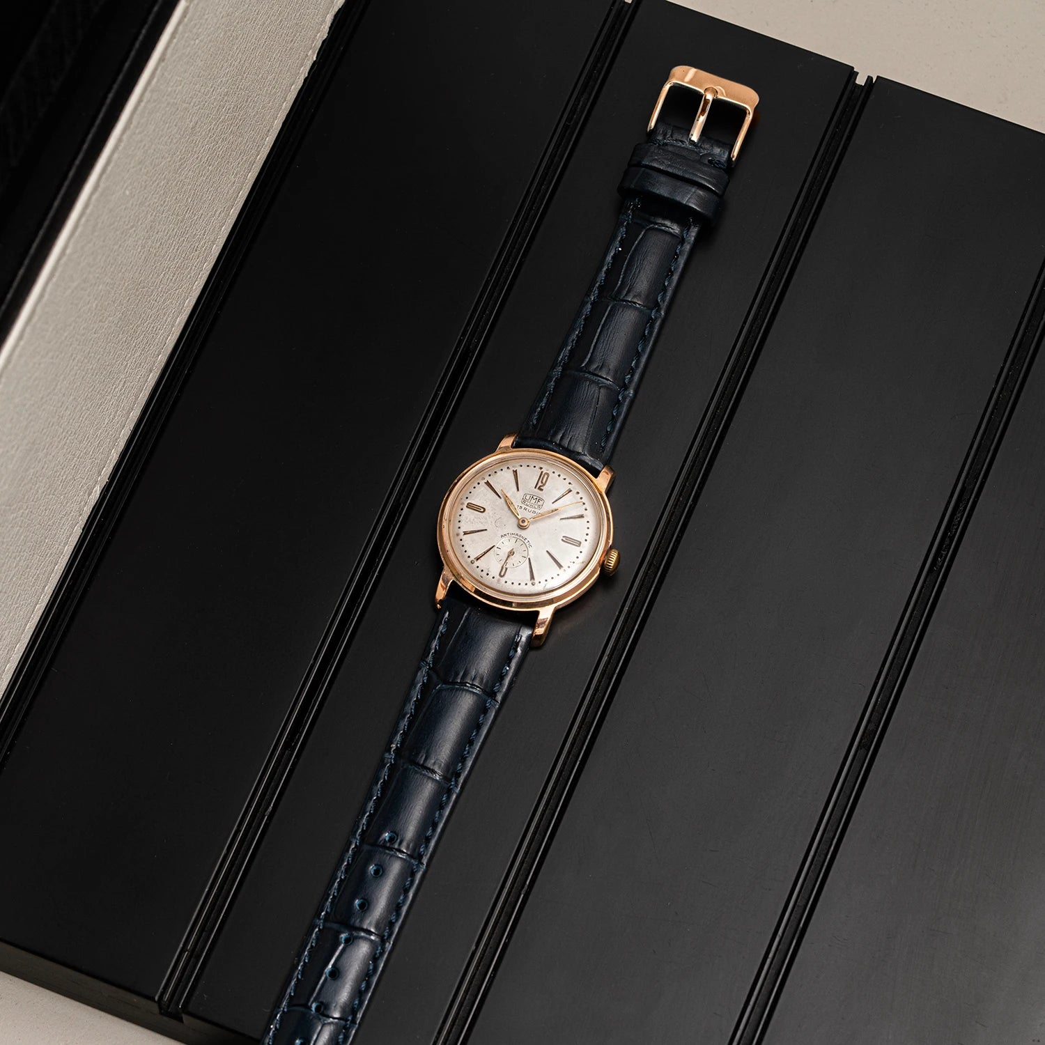 A luxury wristwatch UMF Ruhla, Cal. UMF 2 with a rose gold case, white dial, and dark blue alligator leather strap, presented diagonally on a black textured box.
