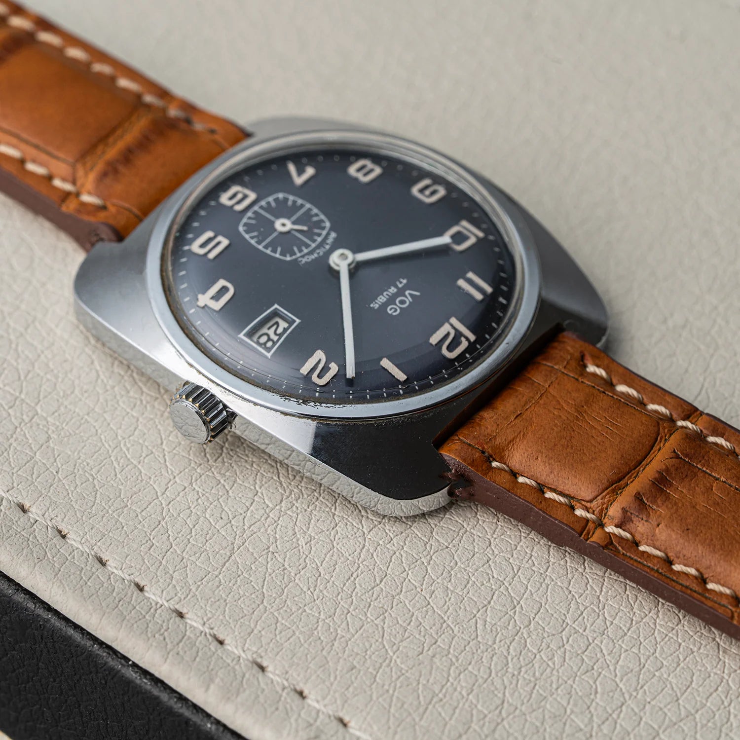 A close-up of a vintage-style wristwatch Vog 17 with a dark gray dial, silver-tone case, and a brown leather strap with white stitching, resting on a textured light beige surface.