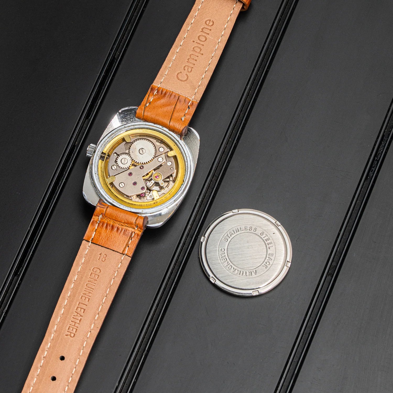 A vintage-style wristwatch with a tan leather strap and exposed mechanical movement, shown on a dark textured background with a separate round metal case placed beside it. The strap is embossed with 'Campiono' and 'Genuine Leather.