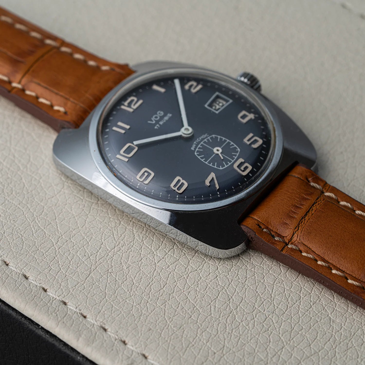 A close-up of a VOG wristwatch with a dark blue dial, silver-toned case, and a brown leather strap, resting on a textured light beige leather surface.
