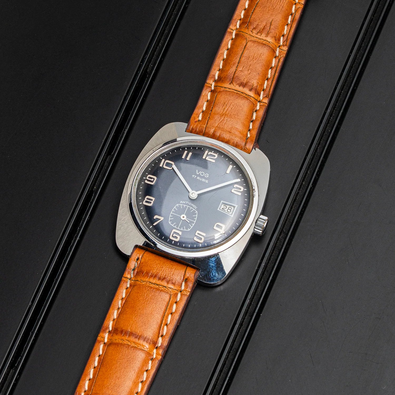A wristwatch Vog 17 with a silver-toned metal case, a dark gray dial, and a brown alligator-patterned leather strap, shown diagonally on a black textured background.