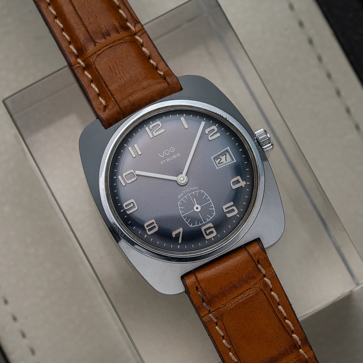 A close-up of a vintage-style wristwatch Vog 17 with a round silver-tone case, dark gray dial, and brown leather strap, resting on a light beige surface.
