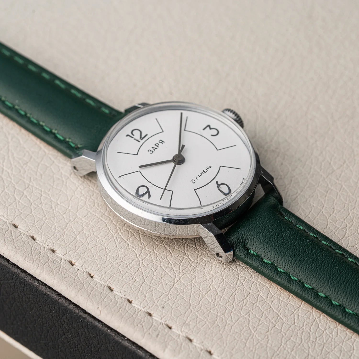 A close-up of a Zaria Z2009B wristwatch with a silver-toned case, white dial, and dark green leather strap, resting on a textured beige leather surface.