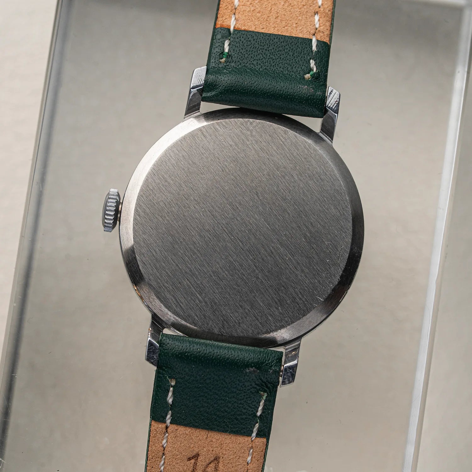 Close-up of the back of a wristwatch Zaria Z2009B with a brushed metal case and a leather strap in dark green and tan, shown against a neutral light gray background.
