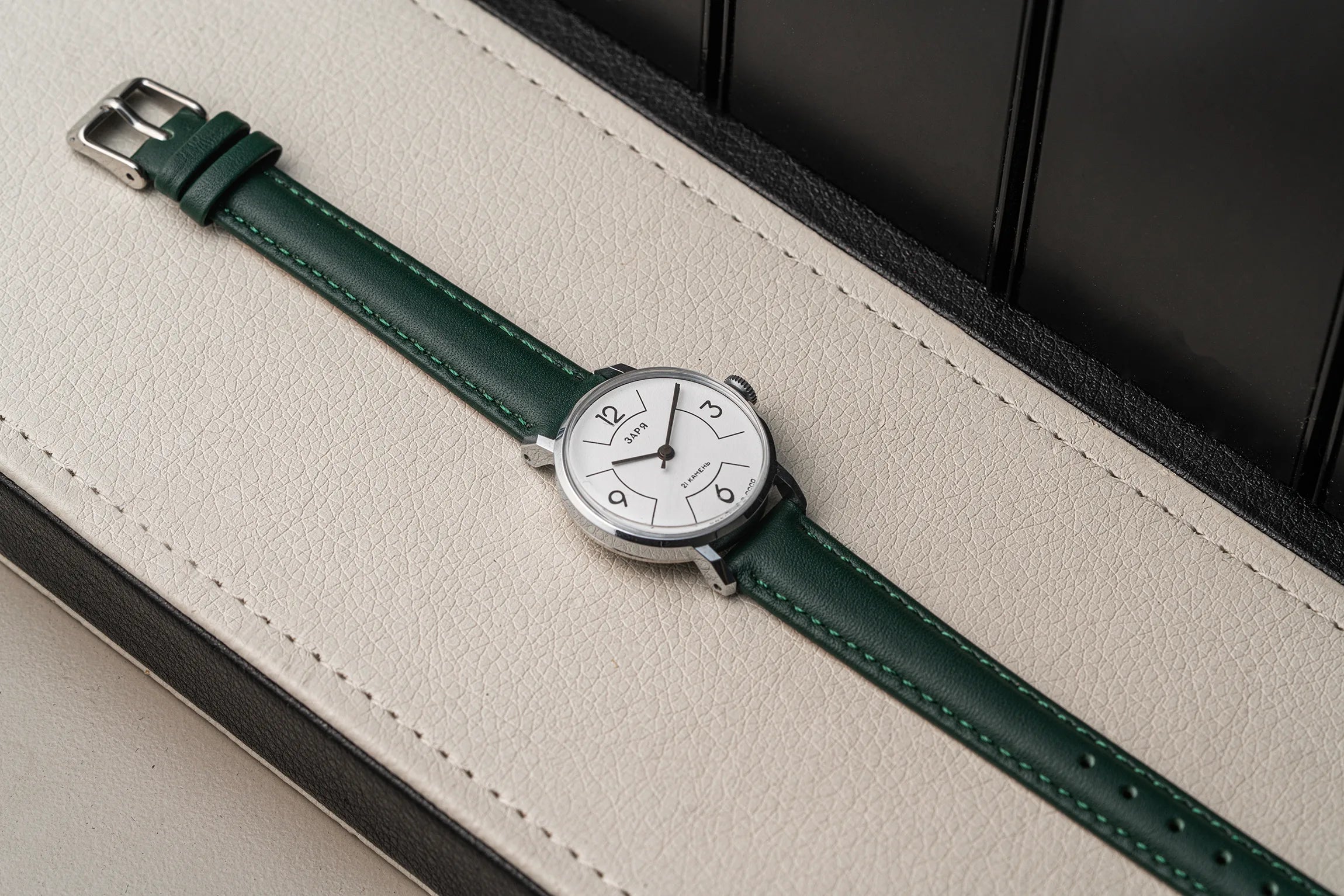 A wristwatch Zaria Z2009B with a dark green leather strap and a silver-toned oval dial, resting diagonally on a cream-colored textured leather surface.