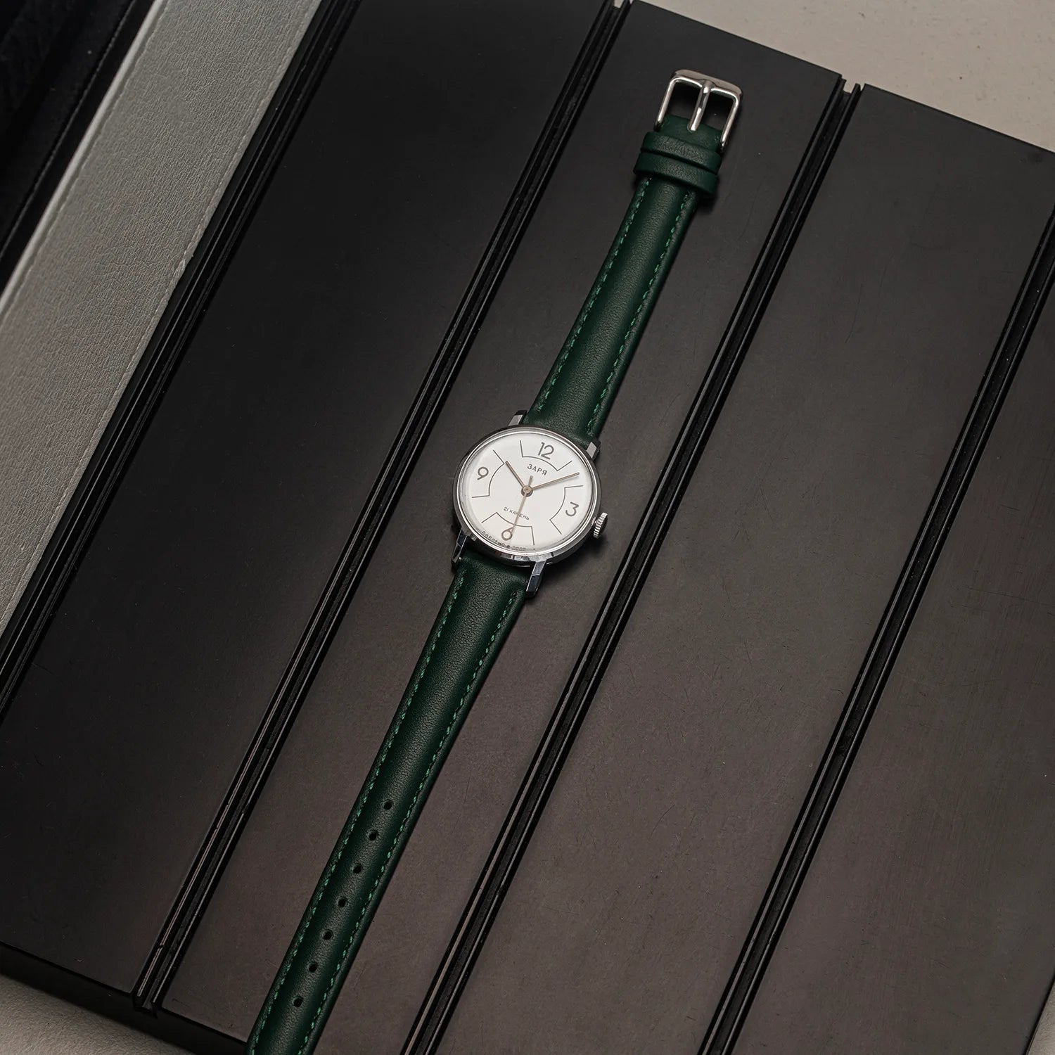 A wristwatch Zaria Z2009B with a silver-toned case, white dial, and black-and-gray striped nylon strap, displayed diagonally on a dark textured surface.