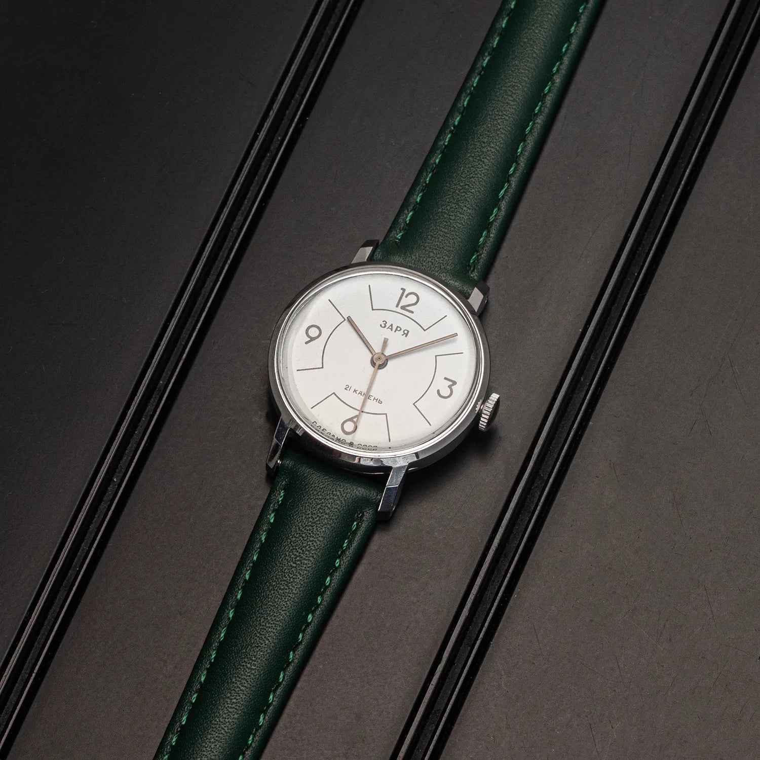 A vintage-style wristwatch Zaria Z2009B with a white dial and a dark green leather strap, resting diagonally on a dark textured surface.